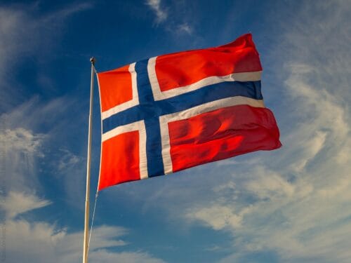 Solen skinner på norsk flagg som friskt blafrer i vinden, mot blå himmel med skyer.